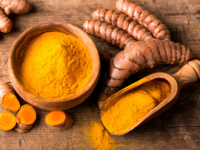 A wooden bowl full of ground turmeric powder sits amongst a pile of chopped turmeric roots